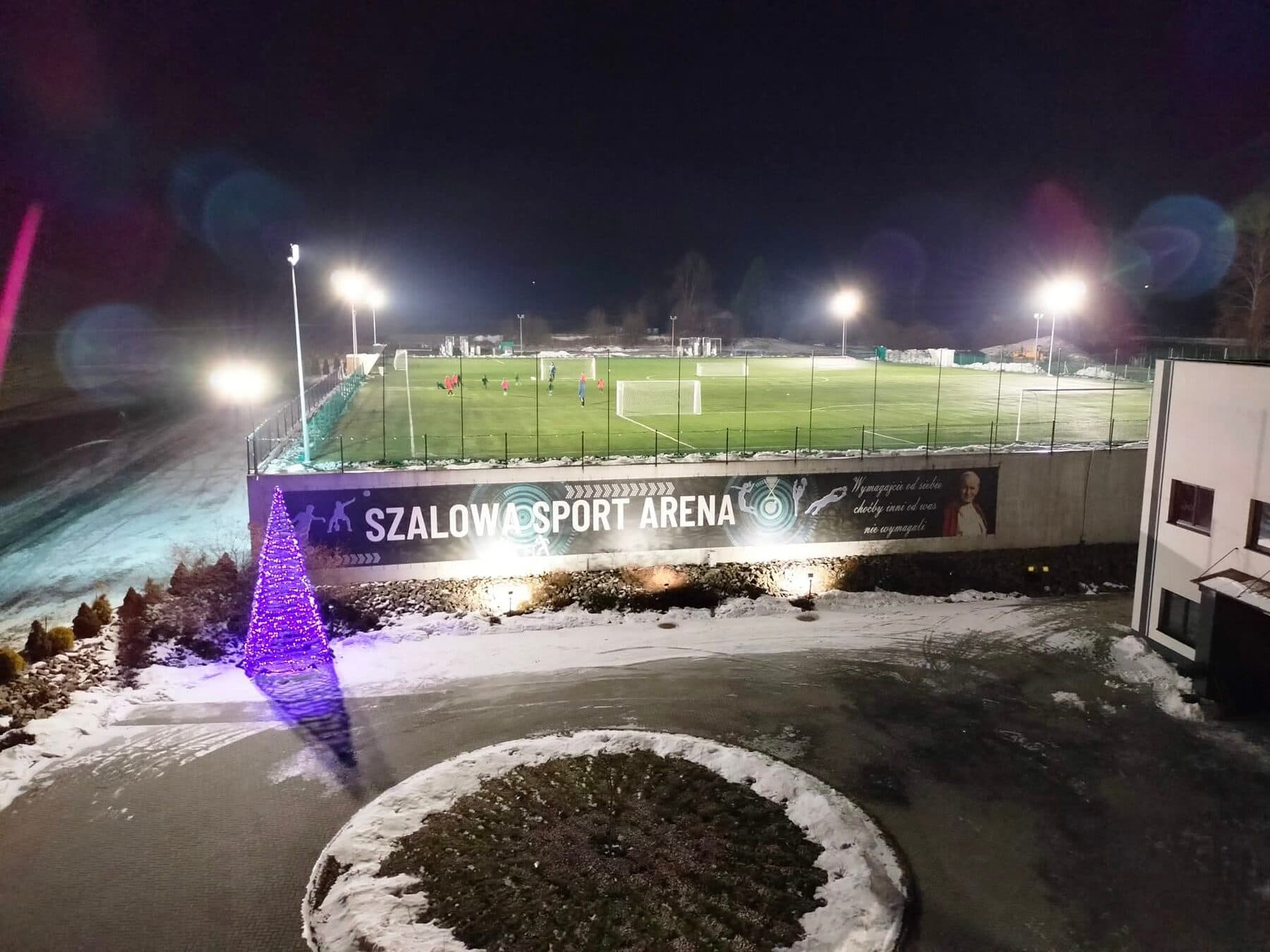 Oświetlone boiska Szalowa Sport Arena po zmroku, z treningiem na murawie i banerem obiektu na murze oporowym