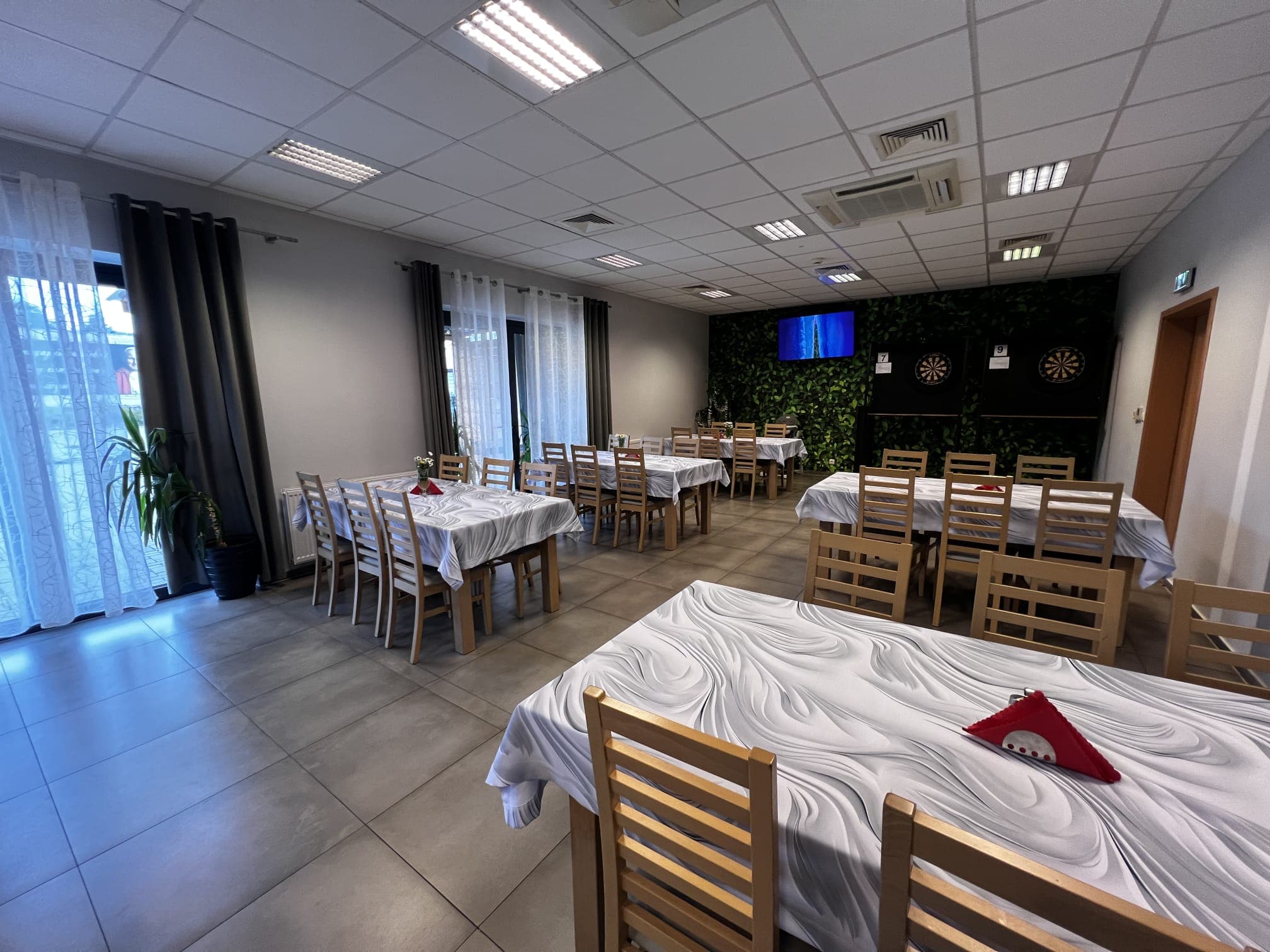 Sala restauracyjna w pełnym ujęciu: rzędy stołów nakrytych białymi obrusami, duże okna z widokiem na taras i ściana z zielenią w głębi